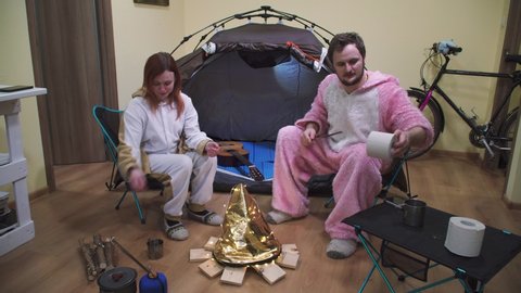 Couple sitting in folding chairs by the tent and bonfire. Camping in the apartment. Adventures at home, traveling around the room. quarantine entertainment Funny, fried marshmallows, toilet paper