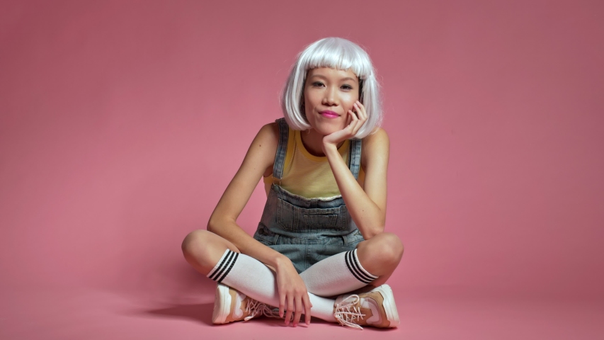 Young positive asian girl sit isolated over pink wall background in white wig blowing kisses