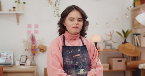 Portrait of beautiful girl with genetic disorder wearing in artists apron and holding paintbrush. Pretty female teen looking and posing to camera while standing in her room