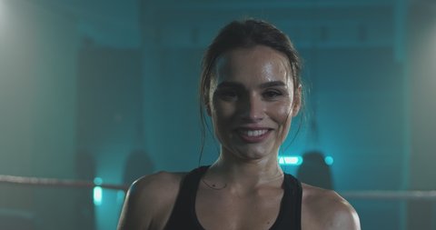 Portrait of beautiful young tired and sweaty woman boxer turning face to camera and smiling cheerfully at dark ring. Close up of pretty girl sportswoman looking at camera with smile after training.