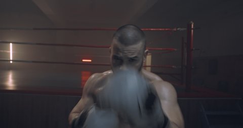 Close up of Caucasian shirtless sweaty strong male boxer in gloves punching with fists to camera at dark ring. Boxing concept. Brutal kickboxer training alone. Sportsman kicking and striking.