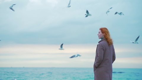 portrait of inspired dream relax, calm one 
beauty redhead girl stands on the pier by the sea, on the coast, by the ocean. winter or autumn, cold weather, birds and seagulls flying. wearing a coat