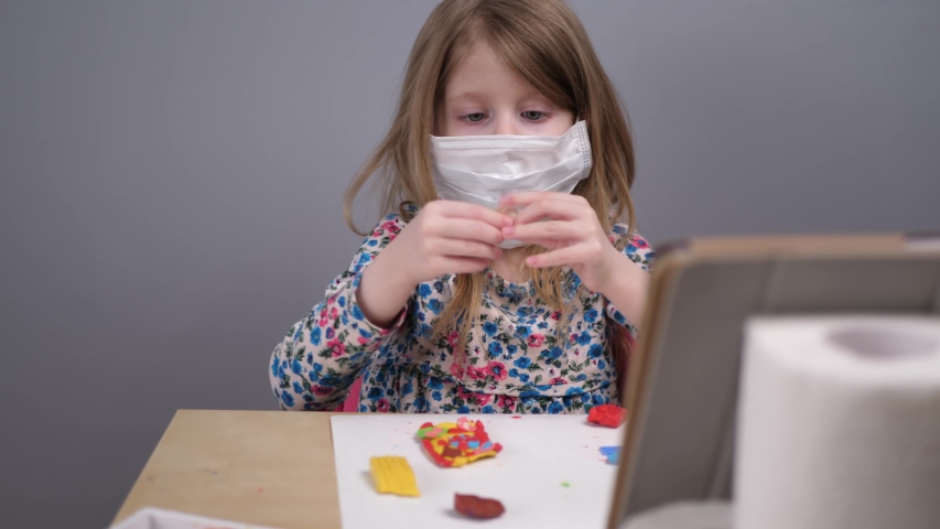 The child attentively watches the lesson on a digital tablet, which is propped against toilet paper rolls. Little girl in a protective face mask sculpts a blood model in an online lesson.