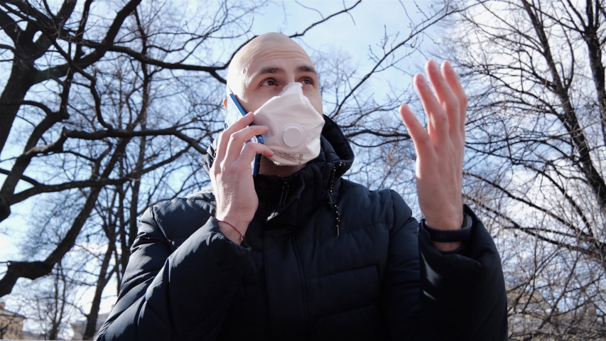 A young Caucasian guy with a bald head who has an infection is sitting in a respirator in a Park in the fresh air and discussing something on the phone