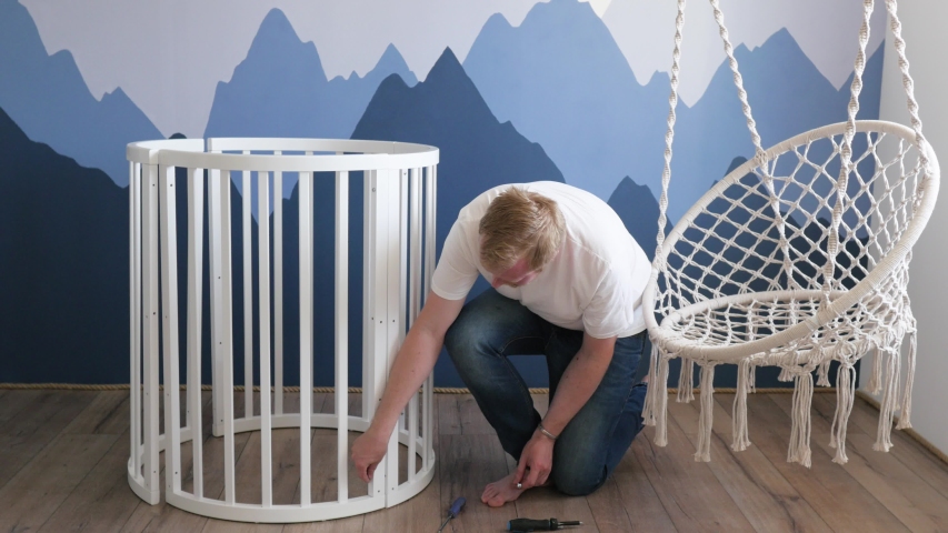 young man collects a crib with a screwdriver for an unborn child. man collects a crib against the background of a wall with mountains. Scandinavian style