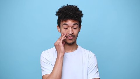 Unshaven boring african american young guy 20s in white t-shirt curly hair posing isolated on blue background studio portrait. People sincere emotions lifestyle concept. Looking at camera rubs his eye