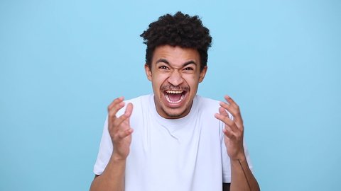 Angry mad african american young guy 20s in white t-shirt isolated on blue background studio portrait. People sincere emotions lifestyle concept. Look at camera screaming scolding protest waving hands