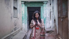 A movement shot of a happy smiling traditional Indian woman girl female walking in salwar kameez in a small narrow lane alley using a mobile phone smartphone cellphone to type a text message