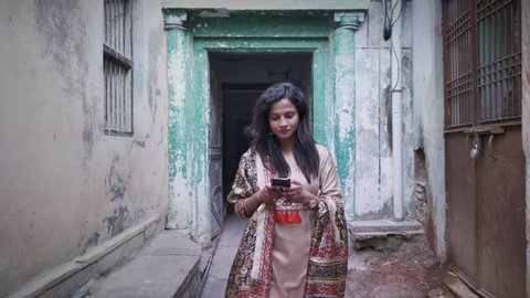 A movement shot of a happy smiling traditional Indian woman girl female walking in salwar kameez in a small narrow lane alley using a mobile phone smartphone cellphone to type a text message