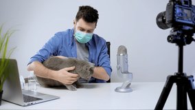 Male Blogger Streaming Wearing Medical Mask Playing with Cat. Video Content Maker. Virus Concept Video. Blogger Medical Mask Use. Man playing with Pet
