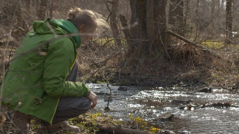 Young handsome woman sitting on the river bank. She picks the first spring flower - coltsfoot and smells it. Water in the river glistens in the sun. Eco friendly concept.