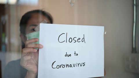 Asian business owner woman wearing face mask or small shop manager attaching business closed sign at shop entrance due to financial crisis from coronavirus covid-19 epidemic outbreak over the world.
