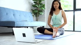 20s young Asian woman in sportswear doing stretching exercises while watching yoga training class on computer laptop online. Healthy girl exercising in living room with sofa couch in the background..