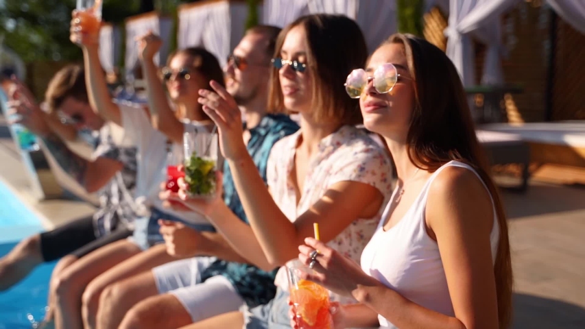 Pretty woman in stylish sunglasses toasting with friends, drinking cocktail of plastic straw at pool party. Group of friends having fun clinking glasses with beverage sitting at poolside on sunny day. | Shutterstock HD Video #1051142506