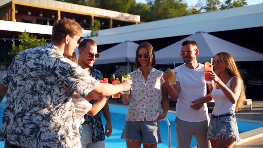 Group of friends having fun at poolside summer party clinking glasses with summer cocktails on sunny day near swimming pool. People toast drinking fresh juice at luxury tropical villa in slow motion. | Shutterstock HD Video #1051142521