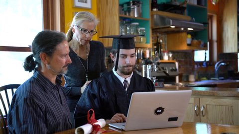 Young man in graduation costume sits in front of computer with family as mother hoods him and happy father looks on during virtual commencement ceremony.