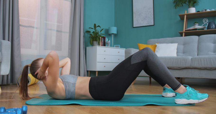 Strong and beautiful athletic fitness girl in sportswear doing press exercises in living room with minimalistic interior. Fitness, home, exercising and diet concept. Healthy and sport lifestyle.