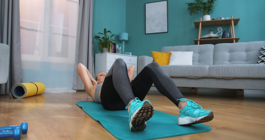 Strong and fit playful girl in grey athletic outfit energetically exercising in bright and spacious living room with minimalistic interior. Fit woman doing abs and press exercises on floor at home