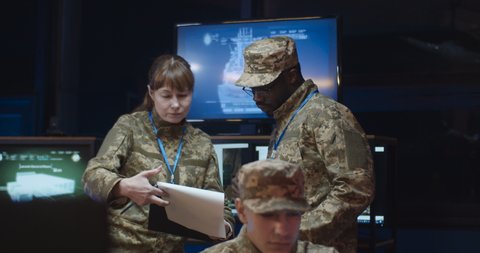 Mixed-races man and woman, officers in camouflage talking and discussing some official data in documents in monitoring room with screens. Male and female army officials working on war strategy.