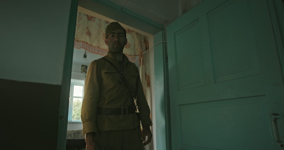 A soldier of the Red Army in World War II, in uniform, stands at the door of an old farmhouse and stares sadly at the camera. Bright sunlight shines through the window.