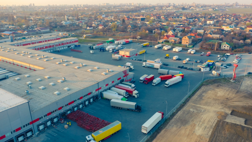 Aerial hyper lapse (hyperlapse - time lapse) of the large logistics park with warehouse, loading hub and semi trucks with cargo trailers standing at the ramps for load/unload goods at sunset Royalty-Free Stock Footage #1051164334