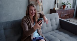 Beautiful young happy Caucasian woman smiles chatting to friends from home using smart phone video app, lying on sofa.