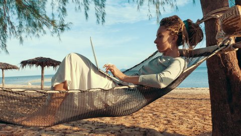 Young woman working on laptop lying in hammock at sand beach of tropical island. Freelance outdoor work concept