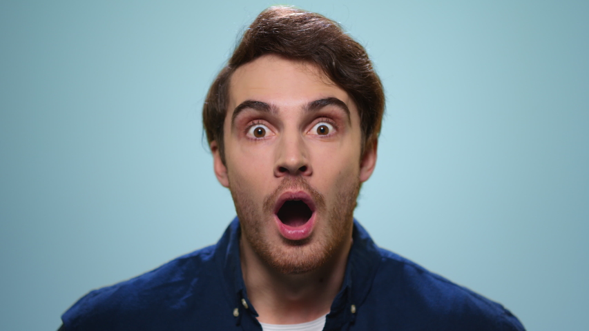 Surprised man looking at camera in slow motion. Handsome guy with wow expression. Close up young man posing on blue background. Portrait of smiling student in studio | Shutterstock HD Video #1051177363