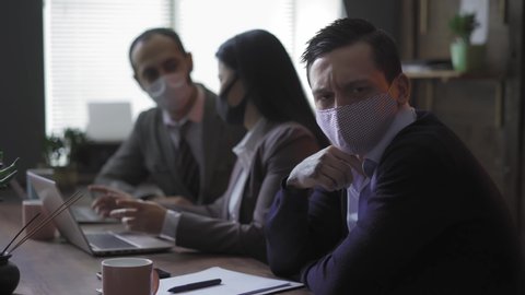 Three businessmen working in quarantine pandemic days sitting in the office and negotiating video calls. Freelancer look at camera on background of two of his colleagues who are talking to each other