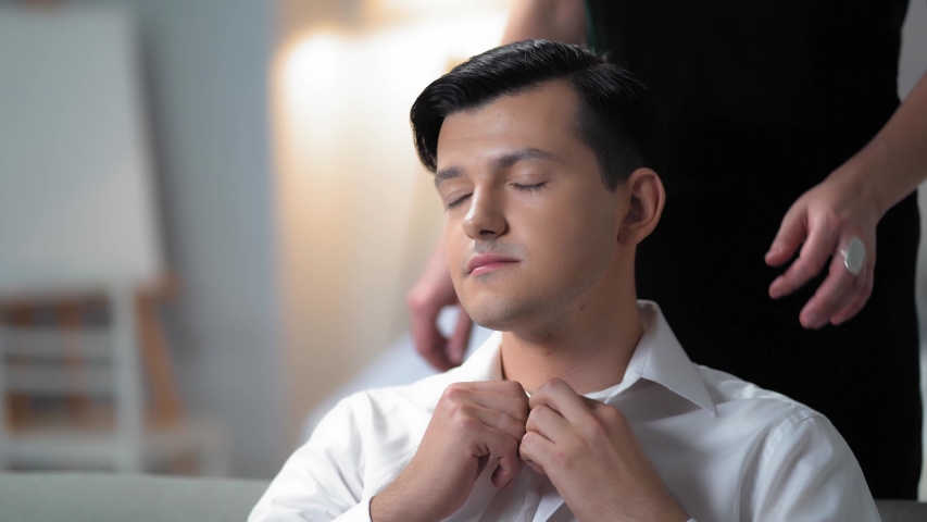 Attractive Young Woman In A Black Dress Is Hugging Gently A Man In A White Shirt Sitting On The Sofa. She Is Caressing Her Boyfriend. The Couple Is In The Stylish Bedroom.