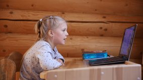 Cute adorable caucasian little blond girl sitting at desk with laptop during online video chat school lesson session with teacher and class. Remote education concept. Self-isolation at quarantine
