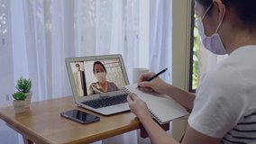 Asian woman wearing mask is working remotely at home by using computer to communicate with make her colleague by video conference during covid-19 or coronavirus infection. social distancing concept