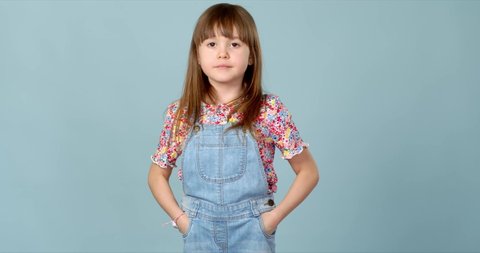Cute little girl standing in dungarees jeans with hands in pockets. Looking at camera with smile. Isolated portrait on blue studio background.