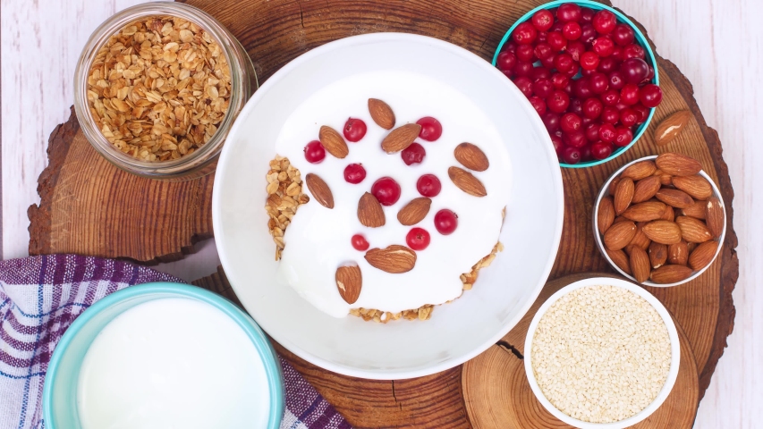Cooking granola with yogurt, almonds, sesame and cranberries. Healthy breakfast concept, white wooden background. Stop motion animation, copy space at the end, top view. | Shutterstock HD Video #1051206385
