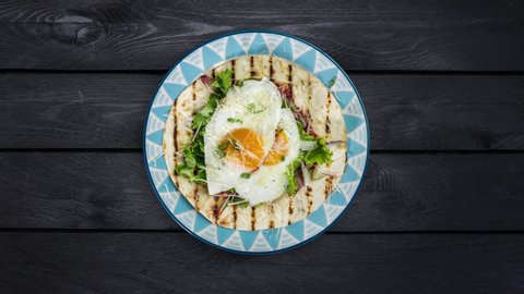 Fried eggs on flour tortilla with green salad and cheese rotating on a plate. Served with cutlery on a black wooden background. Top view