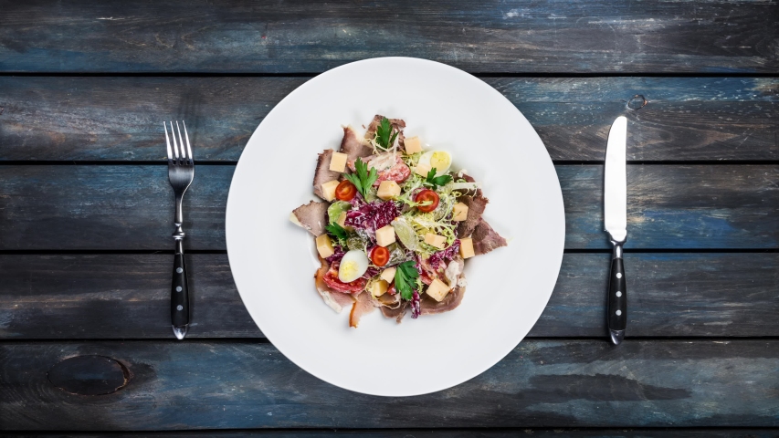 Rotating salad on a plate. Mixed lettuce, tomatoes, turkey smoked, smoked duck, dried horsemeat. Served with cutlery on a colored wooden background. Top view