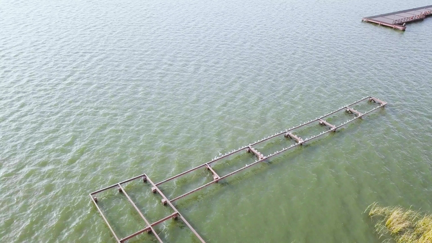 Aerial view of the river with small ripples and the pier under the summer sun. Stock. Flying above green water of the river with small waves and pier construction.