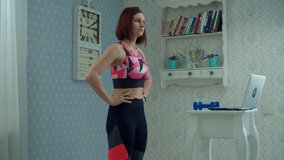 30s woman in sportswear standing in front of laptop and making fitness exercises at home. Female doing sports with dumbbells at home in slow motion. 