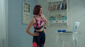 30s woman in sportswear standing in front of laptop and making fitness exercises at home. Female doing sports with dumbbells at home in slow motion. 