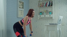 30s woman in sportswear standing in front of laptop and making biceps and triceps exercises with dumbbells at home in slow motion. 