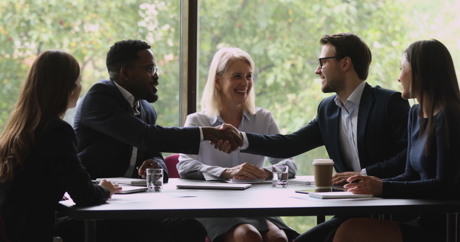 Middle aged smiling businesswoman watching diverse young businessmen in eyeglasses shaking hands after coming to agreement at negotiation meeting. Pleasant business people establishing partnership.