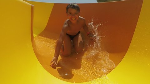 Laughing boy rides down orange slide in a water park. Boy in a swimsuit laughs and slides into the water raising spray
