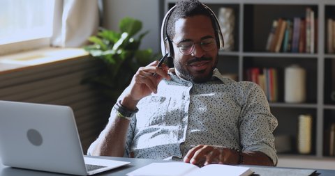 Relaxed young african american guy in glasses wearing headset, enjoying listening favorite audio at pause break time. Happy biracial man beating out music rhythm, resting at table with laptop.