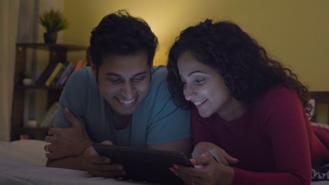 A happy couple husband and wife sleeping on a bed watching a comedy movie web video series online on a digital tablet in a bedroom. A man and woman enjoying a holiday together. Healthy relationship