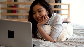 Portrait of happy young Asian girl in casual clothing lying down on bed while making a video call with laptop computer in bedroom at home. Video conferencing technology concept.