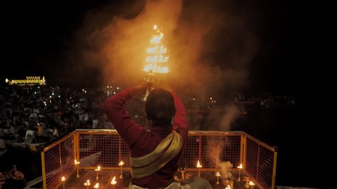 A back shot of a Pujari or Brahmin (Hindu Priest) holding fire lamps or lantern Diyas Performing Ganga Pooja in traditional clothes in the evening at Varanasi ghat, India