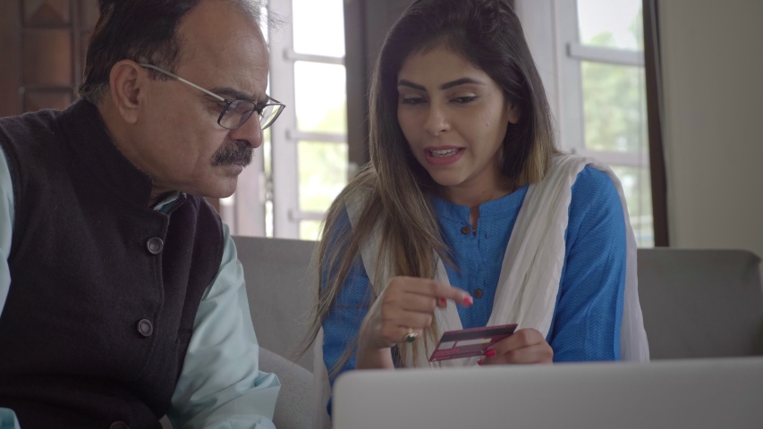 Smiling Daughter teaching her father how to use credit debit card internet banking to make payment on a laptop. A beautiful traditional young woman showing an elderly man how to shop buy online.  Royalty-Free Stock Footage #1051247509