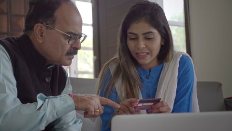 Smiling Daughter teaching her father how to use credit debit card internet banking to make payment on a laptop. A beautiful traditional young woman showing an elderly man how to shop buy online. 