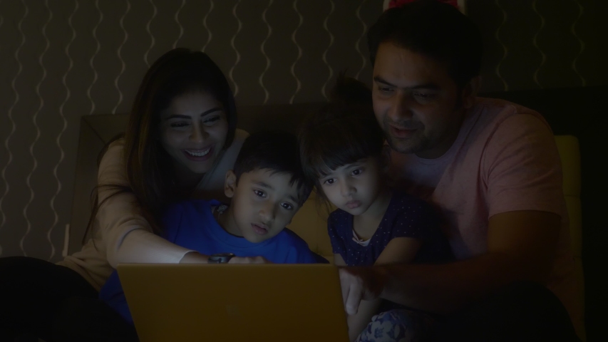A family of four members including parents and two young siblings are enjoying while using tablet or Laptop in the night. A happy family smiling while watching a movie or playing online video game.  Royalty-Free Stock Footage #1051247515