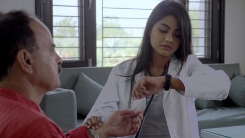 A young female doctor physician checking the heart rate of an old man patient in an interior setup. A medical professional examining taking radial pulse rate by holding the wrist of senior elderly